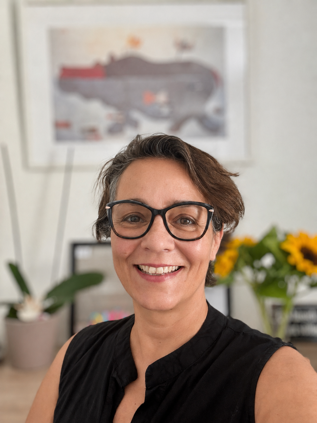 Lächelnde Frau mit Brille im Büro, unscharfer Hintergrund mit Blumen und Wandposter.