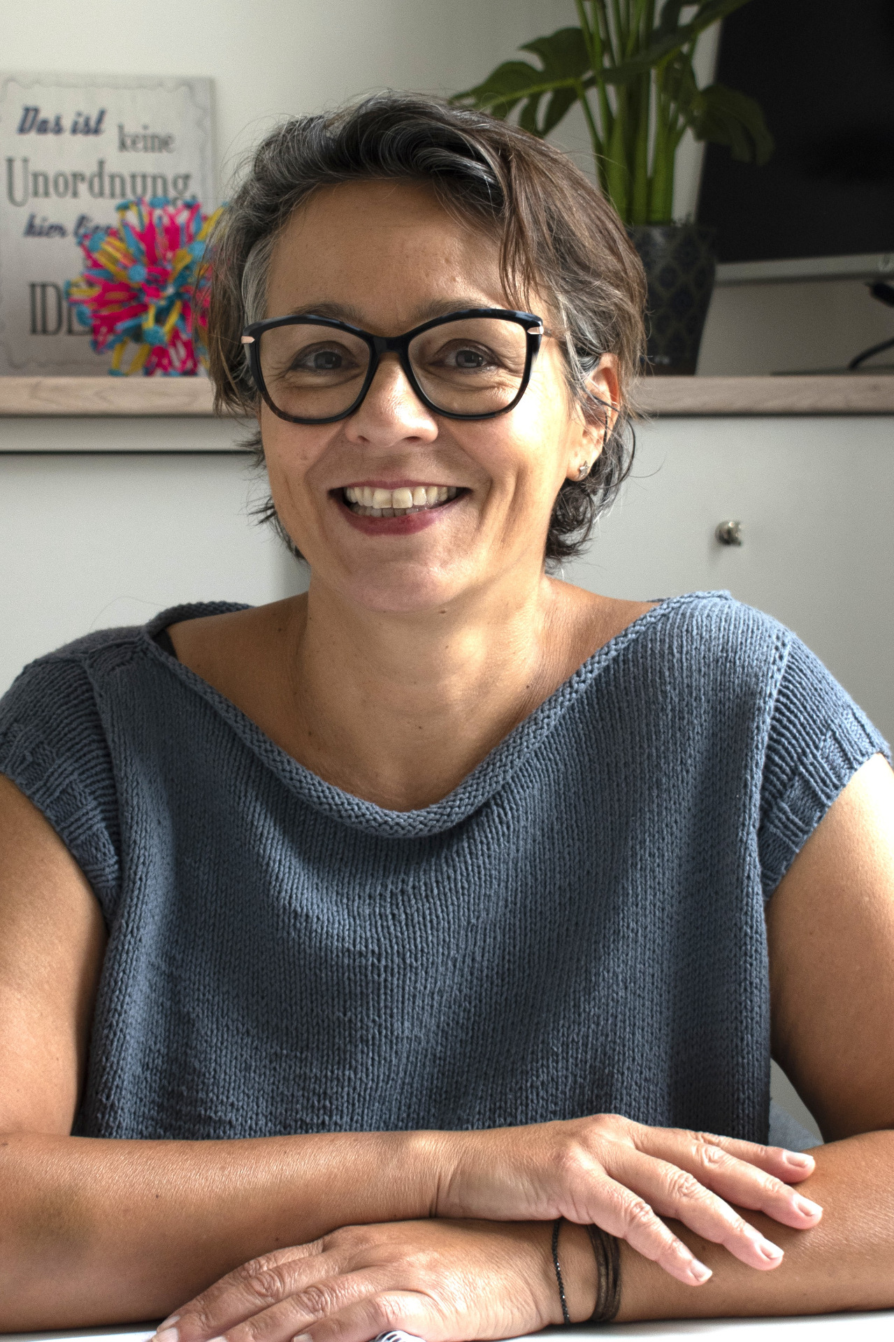 Lächelnde Frau mit Brille im Büro, unscharfer Hintergrund mit Blumen und Wandposter.
